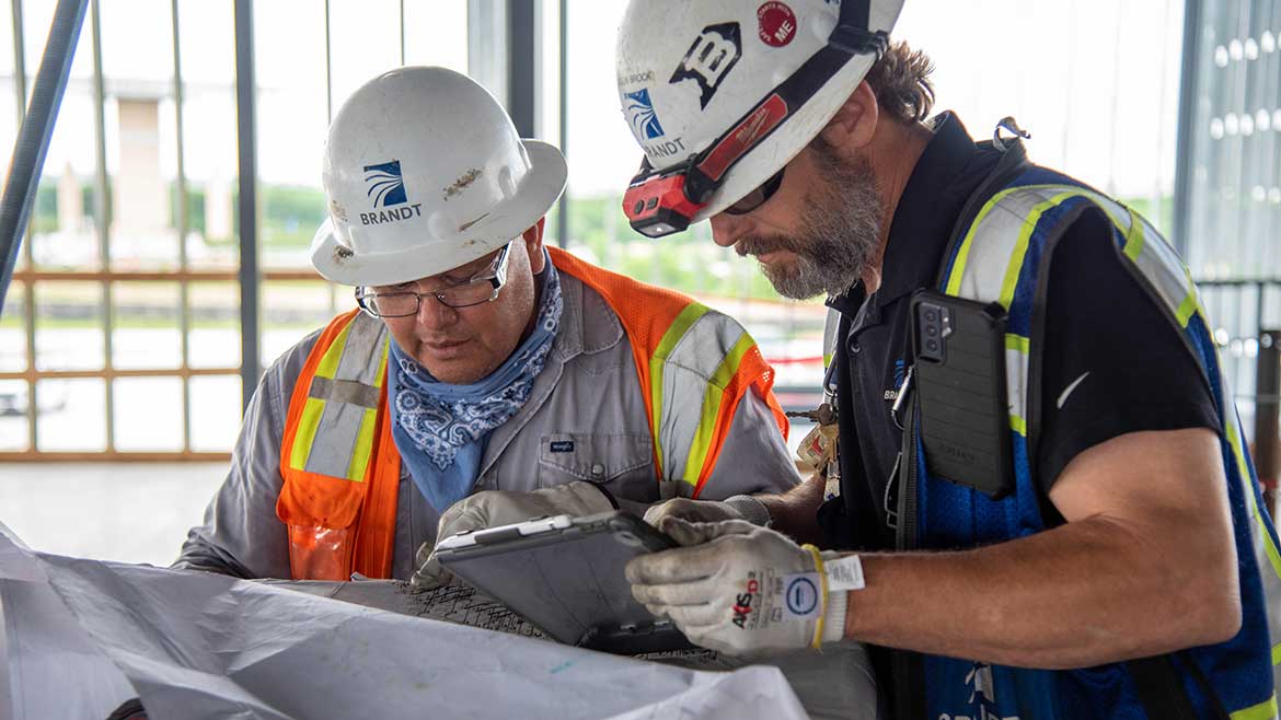 04 PM 1123 PM Profile Brandt Autodesk. Two men in hard hats at construction site looking at plans.